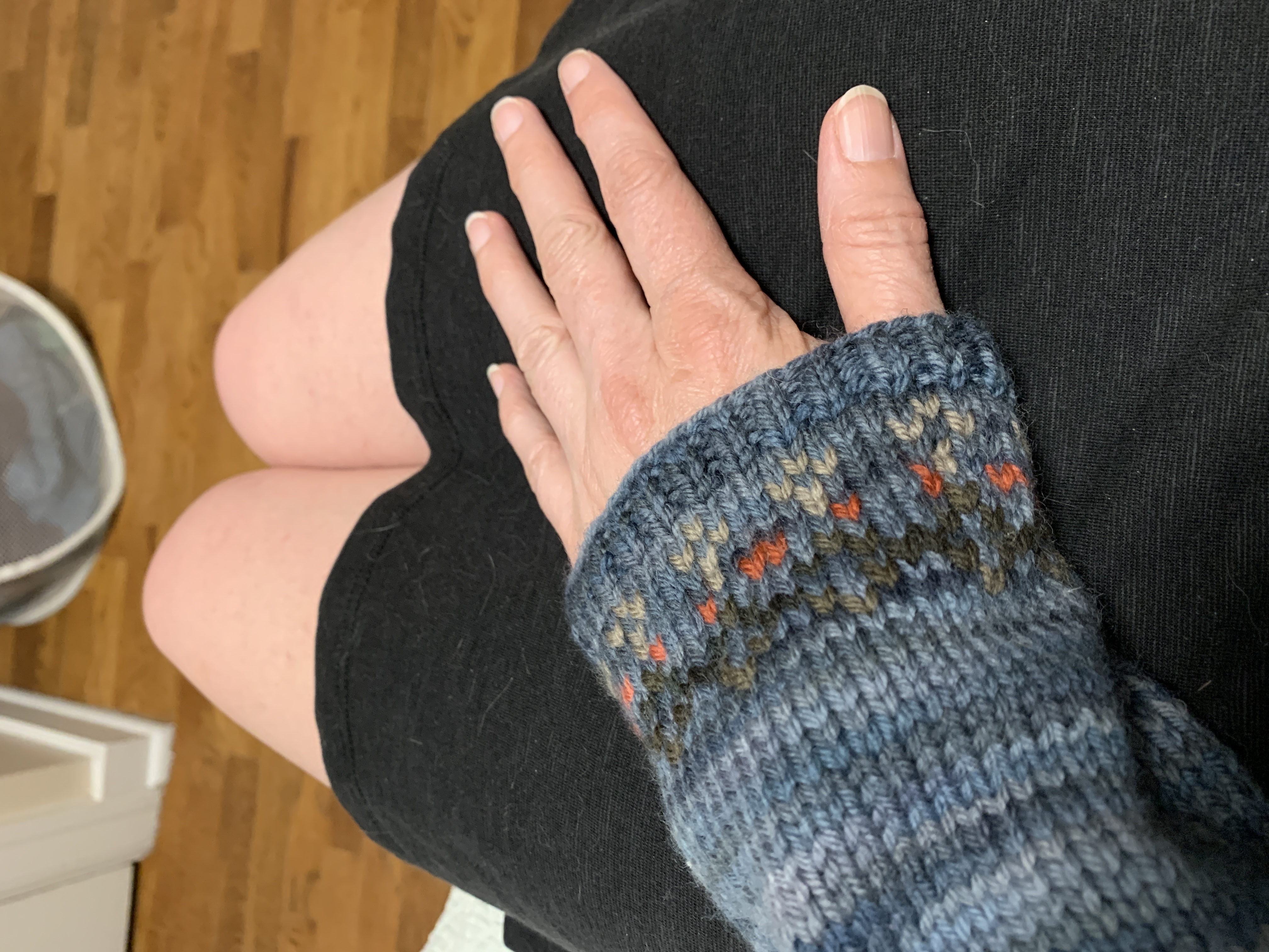 A photograph of someone’s sleeve and light-skinned hand. The sleeve is knitted in shades of blue, with small stitches of tan and orange shaped like hearts close to the hem. 