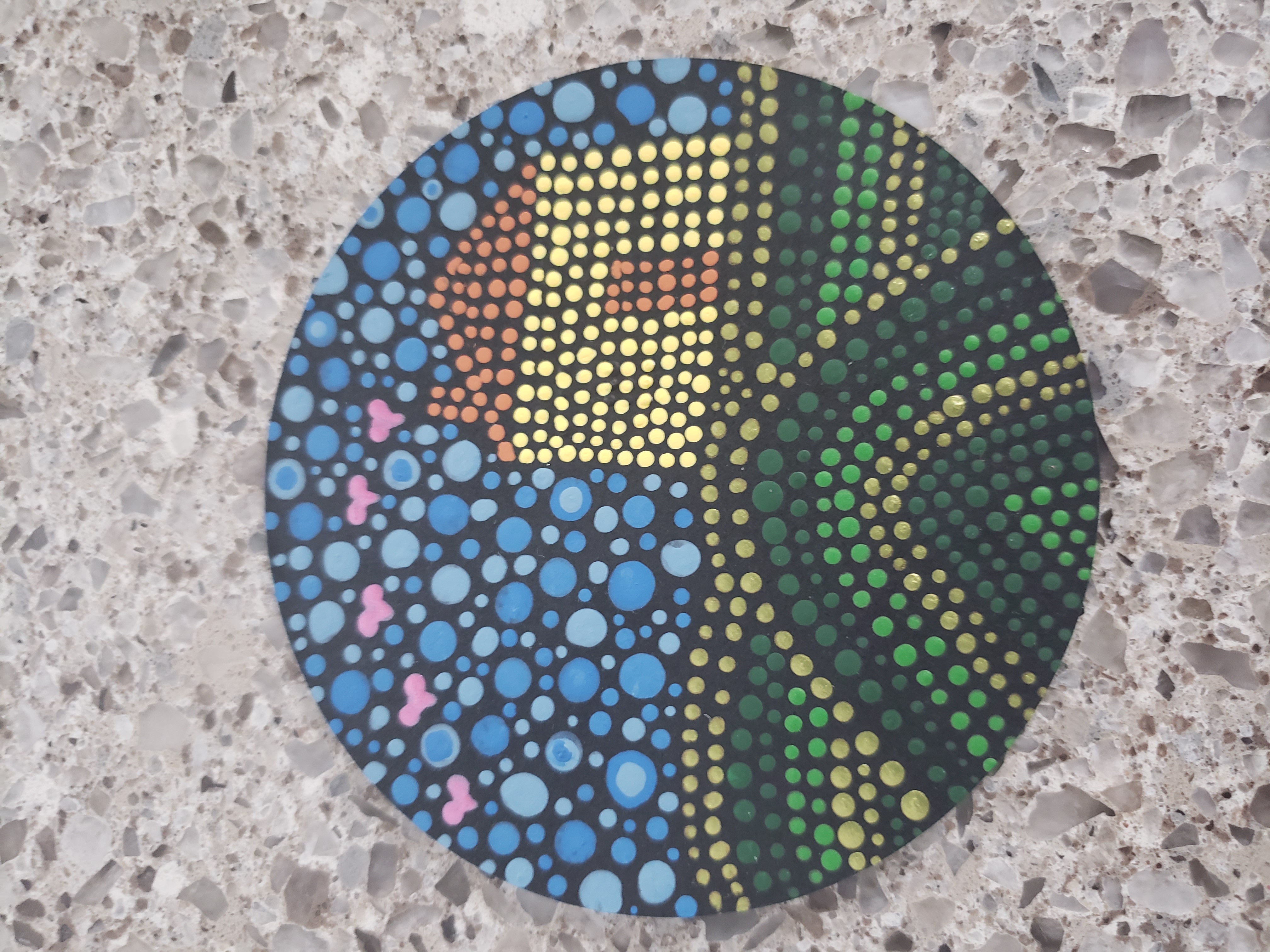 A photograph of an acrylic dot painting on a round disc placed on a tan granite countertop. The background of the painting is black. On top of the black are small painted dots that form the shape of a yellow house on top of three green hills against a blue sky. The hills are formed by lines of dots in varying shades of green. The house is one shade of yellow, with an orange door, chimney and roof. The sky is painted with larger circles in various shades of sky blue. There are five pink hearts in the sky coming from the top of the chimney to the edge of the disc.
