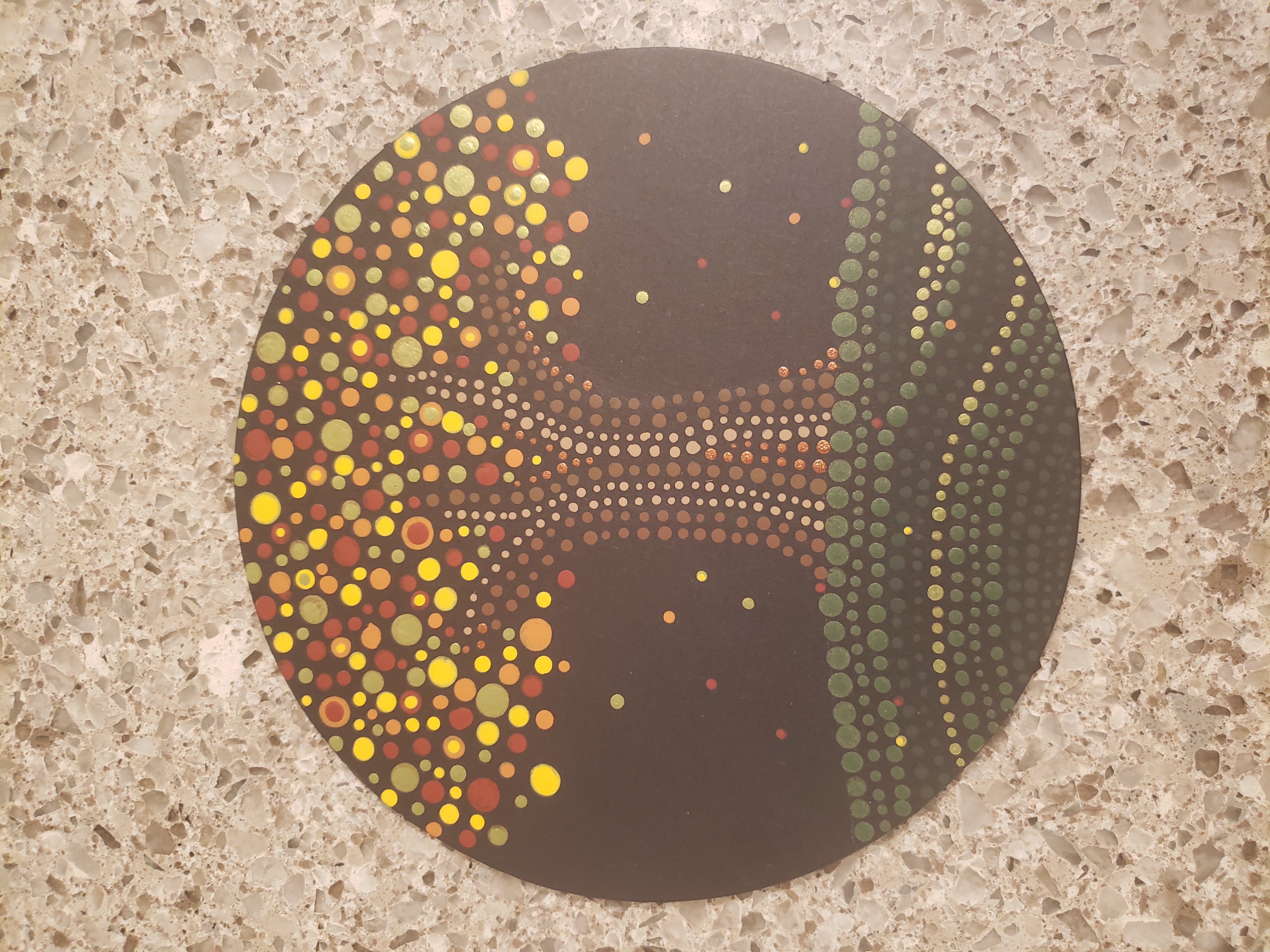 A photograph of an acrylic dot painting on a round disc placed on a tan granite countertop. The background of the painting is black. On top of the black are small painted dots that form the picture of a tree with bright autumn leaves. The tree is on a green hill painted with rows of different colored dots varying from light green at the top to dark green at the bottom of the hill. The trunk and branches of the tree are painted in shades of brown. The dots of the leaves are bigger than the trunk, surrounding the branches in shades of yellow, orange, and red. Smaller dotted leaves fall to the ground.
