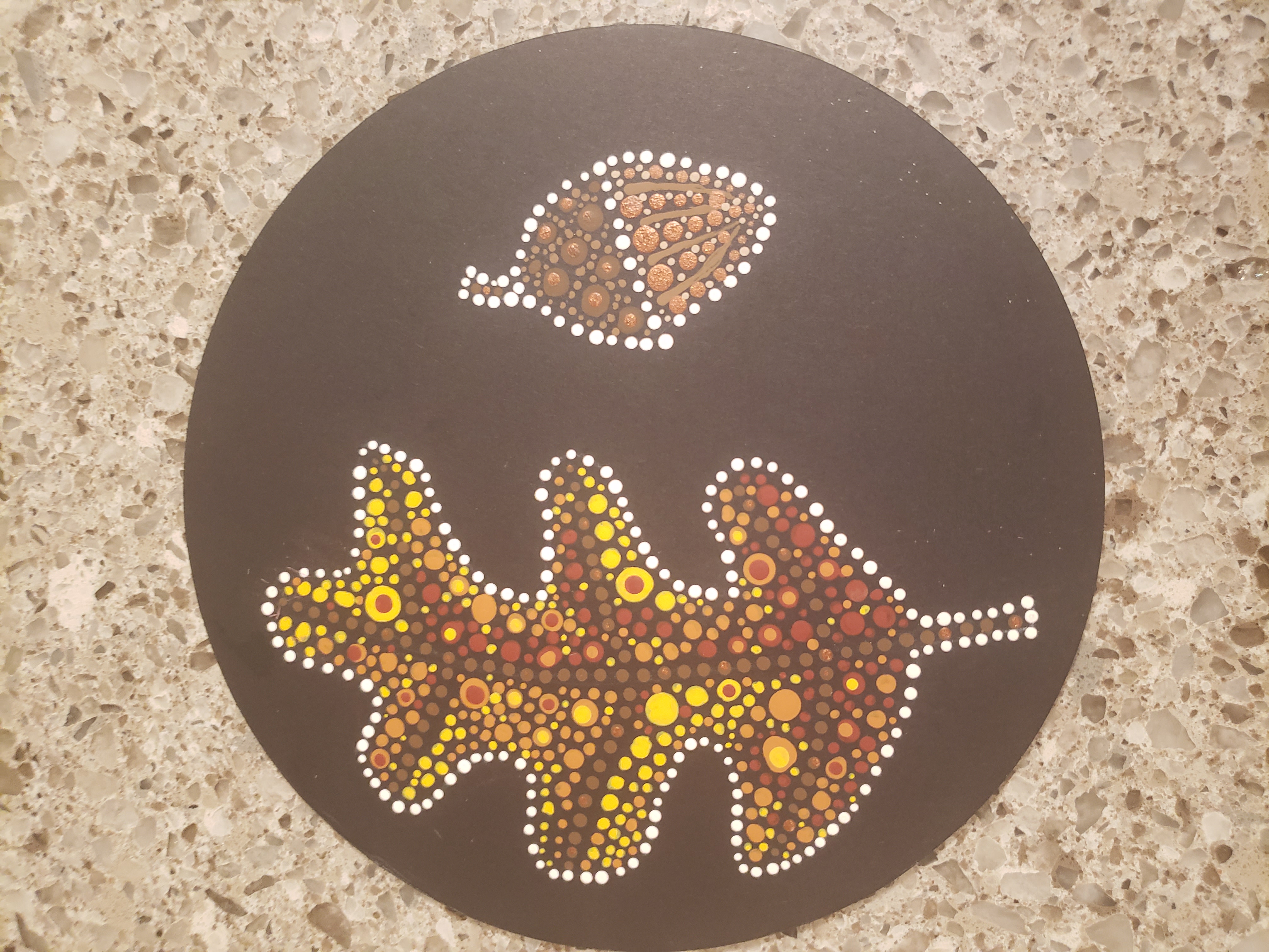 A photograph of an acrylic dot painting on a round disc placed on a tan granite countertop.The background of the painting is black. On top of the black are small painted dots that form the shape of an autumn leaf and an acorn. The leaf is painted in shades of orange, yellow, and red, with brown dotted veins through the center. The acorn is painted in shades of dark brown on the top and light brown on the bottom. Both shapes are bordered with white dots.