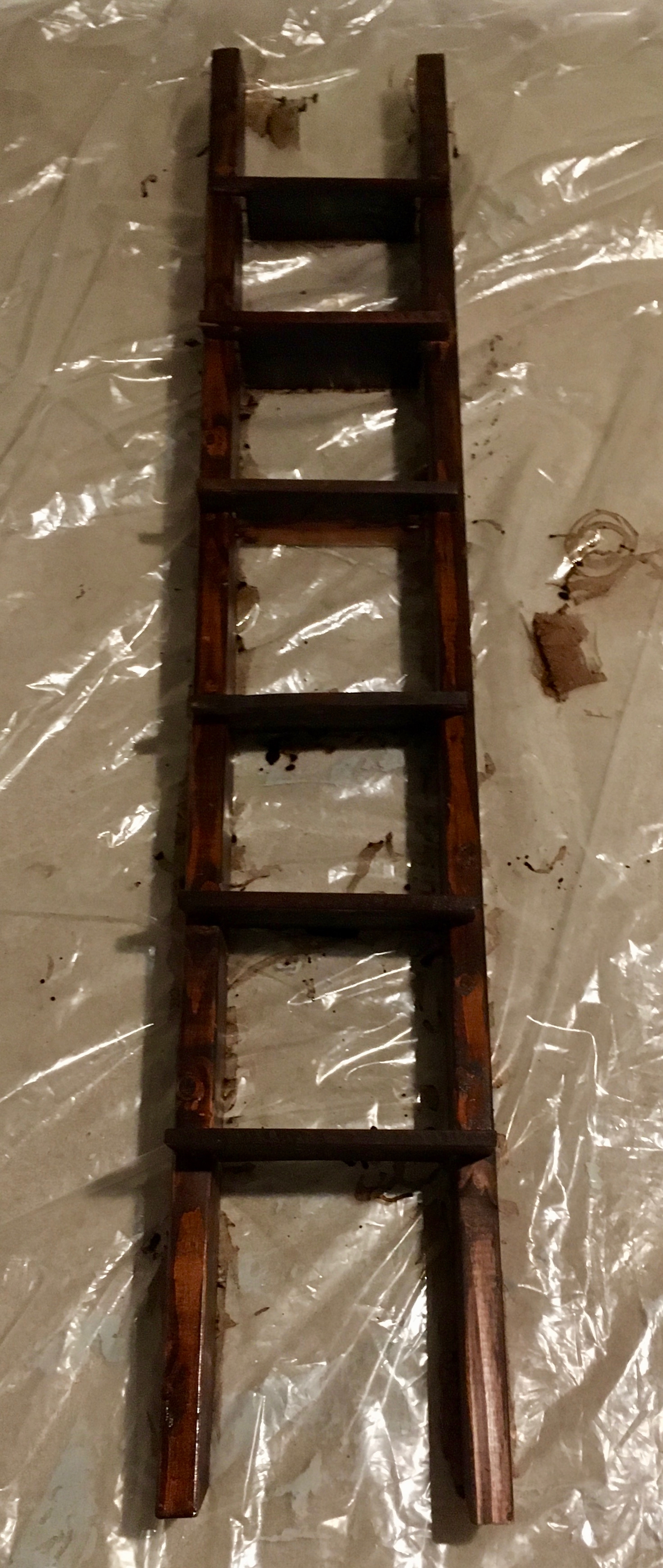 A photograph of a dark brown painted ladder. The ladder is laying on top of plastic as the dark brown paint dries. There are paint smears on the plastic as well as patchiness of the paint on the ladder. The natural color of the ladder is auburn in color and shows through the dark brown paint. A light reflects on the ladder and plastic.