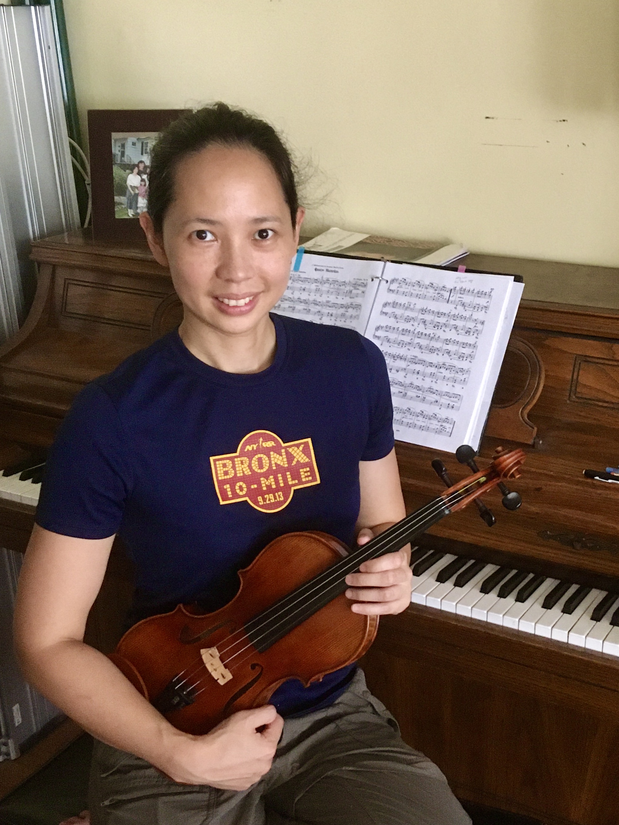 A light-skinned woman sitting in front of an upright piano and holding a violin.
