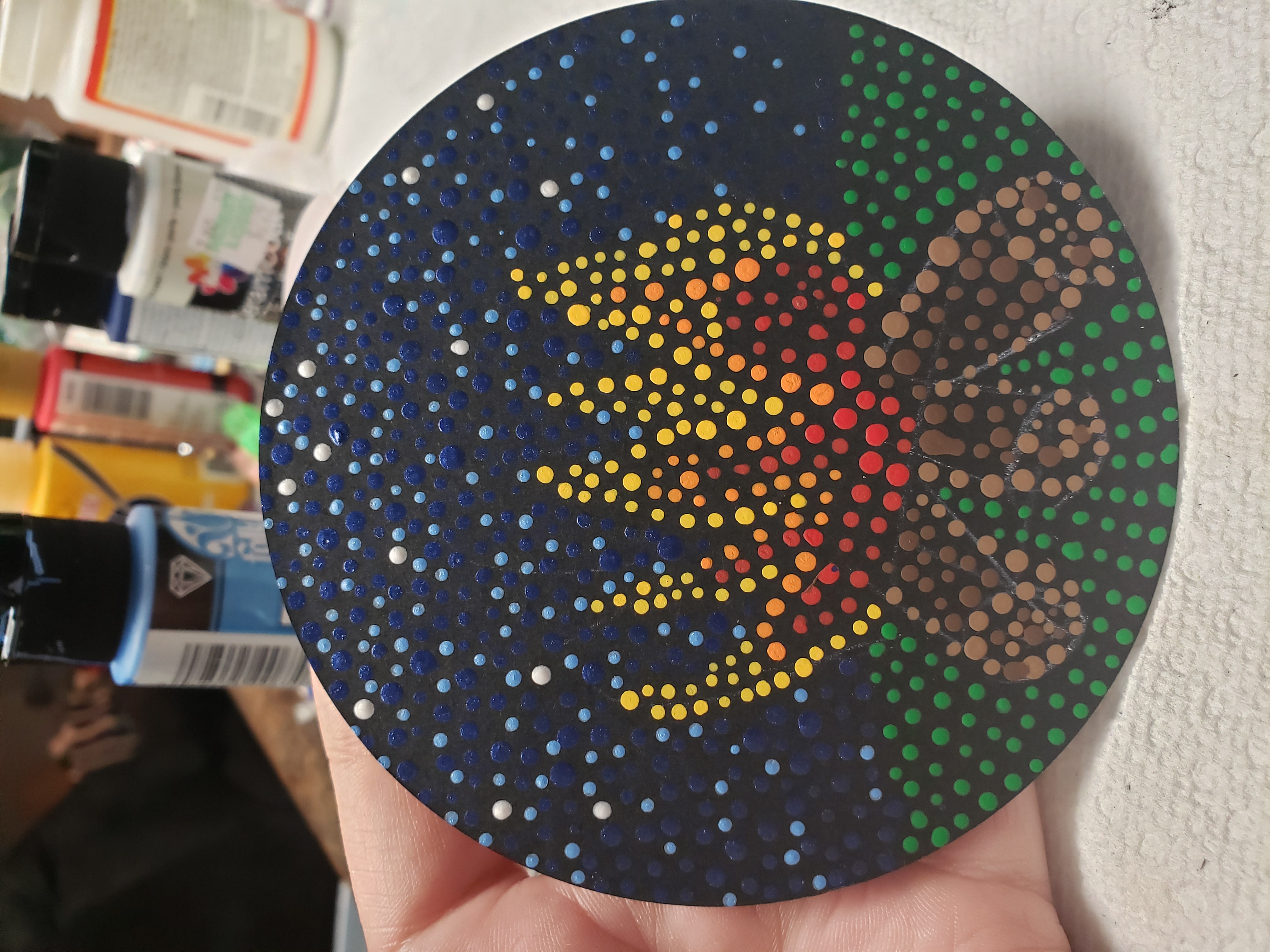 A photograph of an acrylic dot painting on a round disc. The background of the painting is black. On top of the black are small painted dots that form the picture of a campfire on grass underneath a starry sky. The campfire is lit by three logs painted with dots in various shades of brown. The fire starts with red dots above the log that fade to orange and then yellow as the flames rise. Dark blue dots form the night sky, dotted with pale blue and bright white stars.