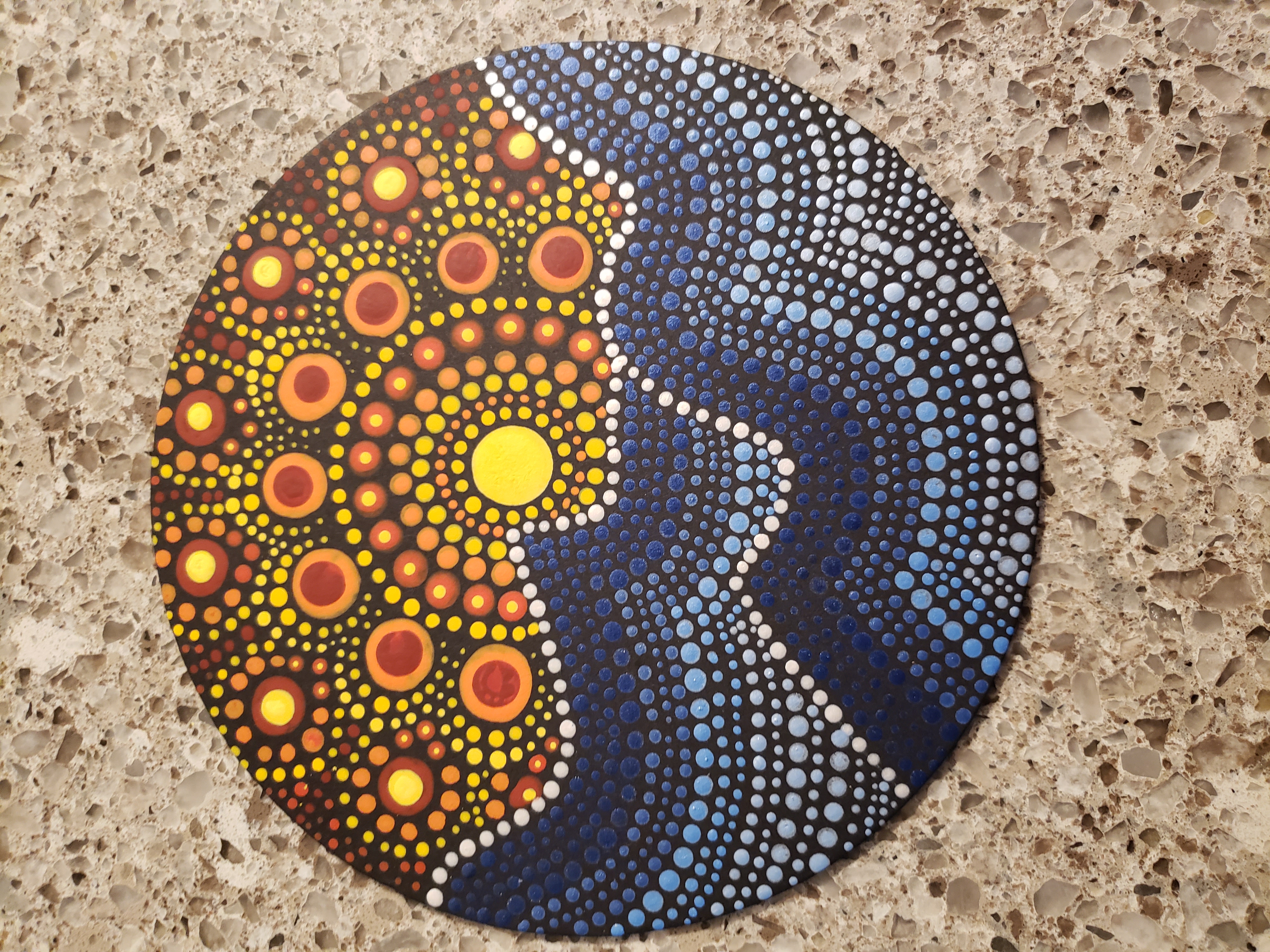 A photograph of an acrylic dot painting on a round disc placed on a tan granite countertop. The background of the painting is black. On top of the black are small dots and circles that form a sun rising over waves. The waves are painted with rows of dots in different colors, starting a deep blue at the crest of the wave and fading to a bluish white at the base. The sunrise is formed by a large yellow dot in the center that is surrounded by an arch-shaped pattern of yellow and orange circles of various sizes.