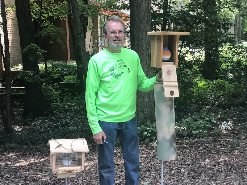 A photograph of a man standing outside in front of trees. The man is white, older, with gray hair, a gray mustache, and a short gray beard. He is wearing glasses, a green long sleeve shirt that says “Autumn classic 8K” surrounded by leaves, and jeans. He is holding a screwdriver. To his left is a bird feeder. To his right is a wooden birdhouse mounted on a pole with a camouflage covering. The bird house is rectangular, with a flat roof and a door that opens at the bottom. Inside the bird house is a toy stuffed bluebird.
