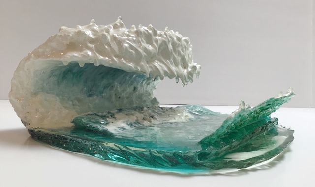 A photograph of a resin sculpture shaped like a cresting ocean wave, on a white backdrop, viewed horizontally from the front facing left with the wave cresting to the right. The base of the sculpture is the semi-transparent blue-green of the ocean. White swirls coat the top of it like swirling sea foam. The wave is on the left side, rising from the base and curling over in the shape of a letter C, with the top of the wave over the ocean base. 
