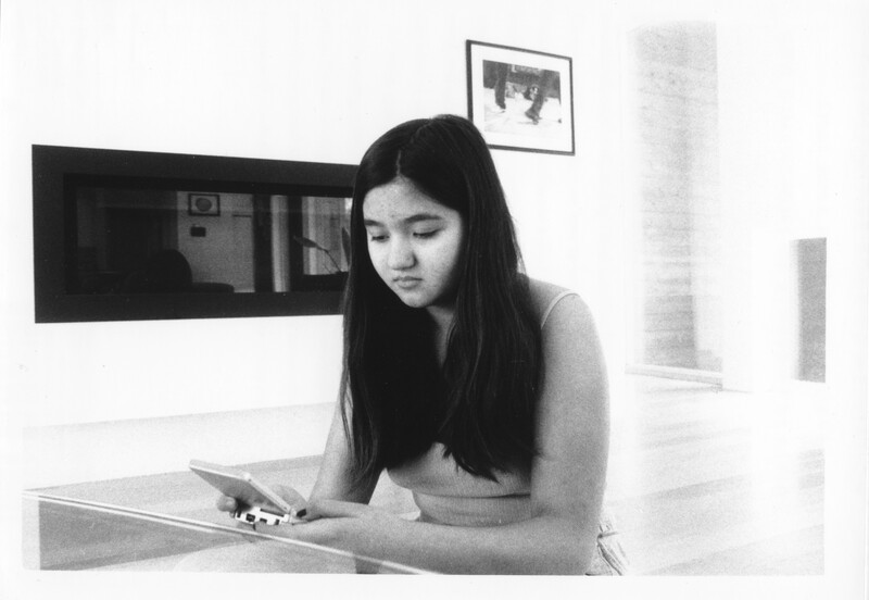 A black and white film-processed photograph of a person playing a handheld video game. The person is shown from the waist up. They have light skin, long dark hair past their shoulders, and are wearing a sleeveless shirt. The room they are in is bright and out of focus, except for a black rectangular built-in fireplace just over their left shoulder. The surface of the fireplace reflects the room. The photograph has a slightly grainy texture.