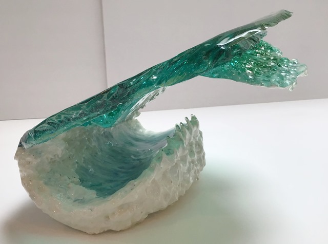 A photograph of a resin sculpture shaped like a cresting ocean wave, on a white backdrop, viewed horizontally from the side and placed upside down with the wave cresting to the right. The base of the sculpture is the semi-transparent blue-green of the ocean. White swirls coat the top of it like swirling sea foam. The wave is on the left, rising from the base and curling over in the shape of a letter C, with the top of the wave coming over the ocean base. 