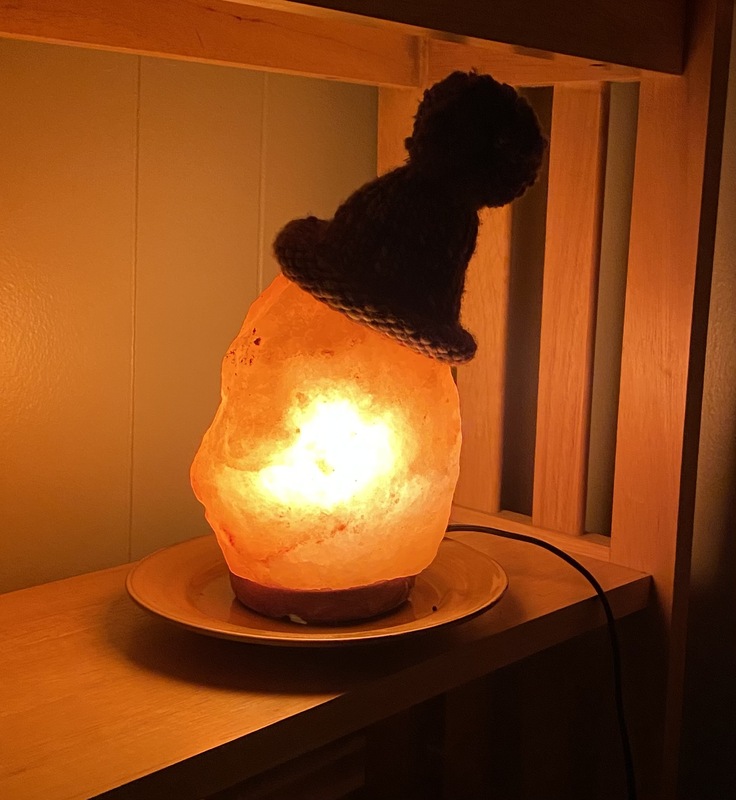 A photograph of a glowing orange salt lamp wearing a knitted hat. The salt lamp takes up the center of the picture, casting a sunset orange glow over the shelf it is on and the wall behind it. The hat is crooked on the top of the lamp, shaped like a triangle, with a pom-pom on top. The brim of the hat is rolled up on itself.