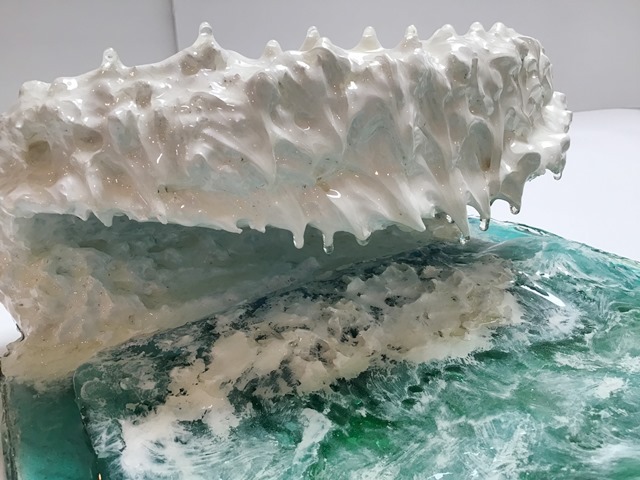 A photograph of a resin sculpture shaped like a cresting ocean wave, on a white backdrop, viewed vertically from the side with the wave cresting towards the viewer. The base of the sculpture is the semi-transparent blue-green of the ocean. White swirls coat the top of it like swirling sea foam. The wave is in the back, rising from the base and curling over in the shape of a letter C, with the top of the wave coming forward over the ocean base. 
