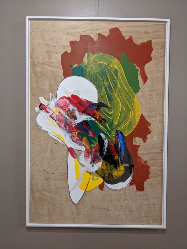 A photograph of an abstract acrylic painting on a wooden block with a white frame. The painting is many different colors swirled together. The edges on the right are red-brown and ragged, and the edges on the left are white and smooth. The center is an oval-shaped patch of green and yellow above an intricate oval of many colors.