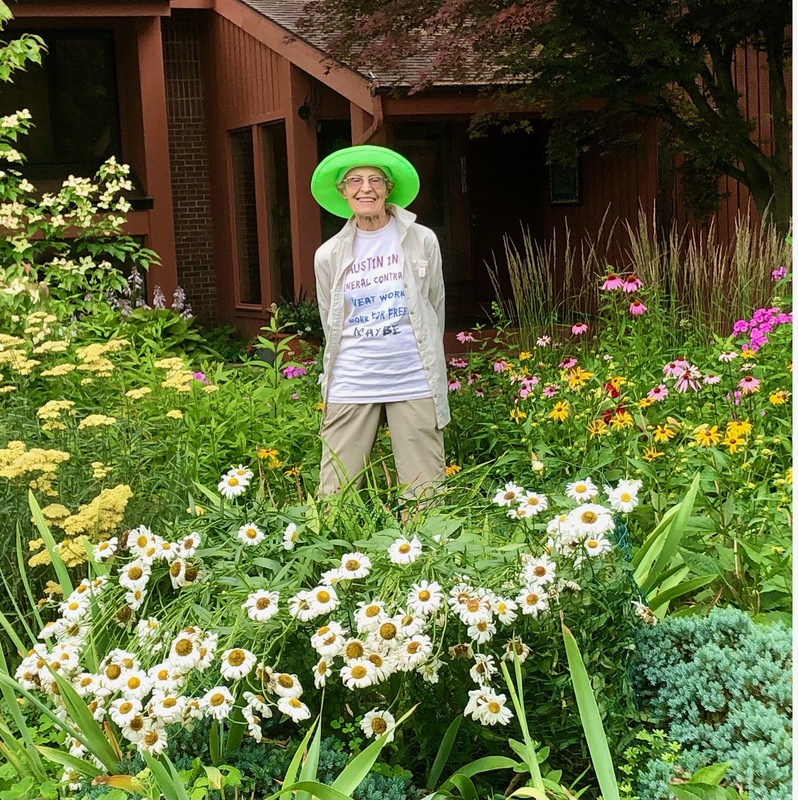 A photograph of a smiling woman standing in the middle of a lush green garden. She is white and elderly. Surrounding her are many different plants and flowers that are taller than her knees. In front of her are white flowers with many petals and yellow centers. On her left are yellow flowers, and on her right are yellow and pink flowers. She is wearing tan pants and a tan jacket over a white t-shirt with text that is unreadable and a bright green sun hat. 