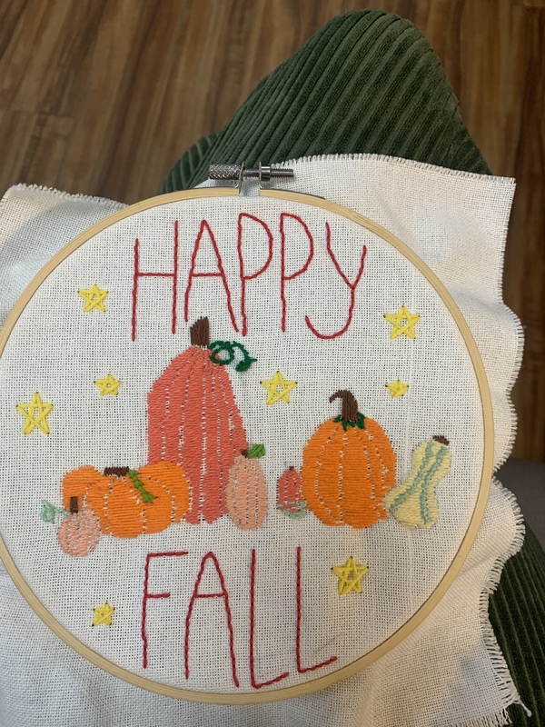 A photograph of embroidery held in place by a circular embroidery hoop. The embroidery reads “happy fall” in red, with “happy” at the top and “fall” at the bottom. In the middle are pumpkins and gourds of various shapes and sizes, colored different shades of orange, with a single yellow and green striped gourd on the right. Yellow stars are scattered around the entire piece. 