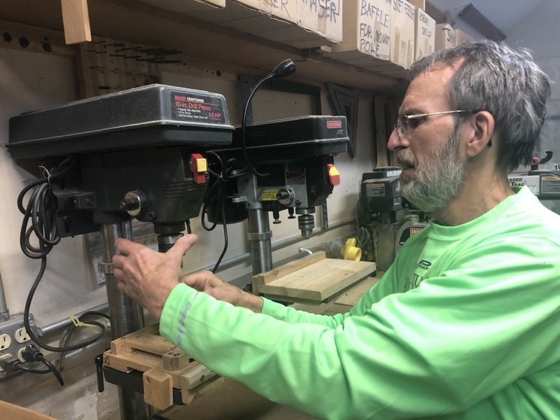 An older white man in a green shirt working on a drill press machine.