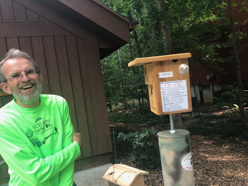 A photograph of a smiling man standing next to a wooden bird house. The man is white, older, with gray hair, a gray mustache, and a short gray beard. He is wearing glasses and a green long sleeve shirt that says “Autumn classic 8K” surrounded by leaves. The bird house is mounted to a pole covered in camouflage that is almost as tall as he is. The bird house is rectangular, with a roof that extends out over it and a tiny circle for the birds to enter from. Below the entrance is a black engraving of a bird. There is a label on the side of the bird house with text that is unreadable.