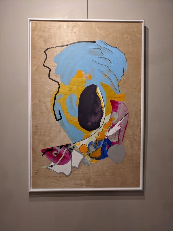 A photograph of an abstract acrylic painting on a wooden block with a white frame. The painting is many colors swirled together in an oval shape with a patch tailing it on the left. The top of the painting is blue. At the center, there is a black oval, surrounded by swirls of gold and blue. The bottom is red on the edges, white in the middle, and orange and blue in the center. Black lines trace the shapes.