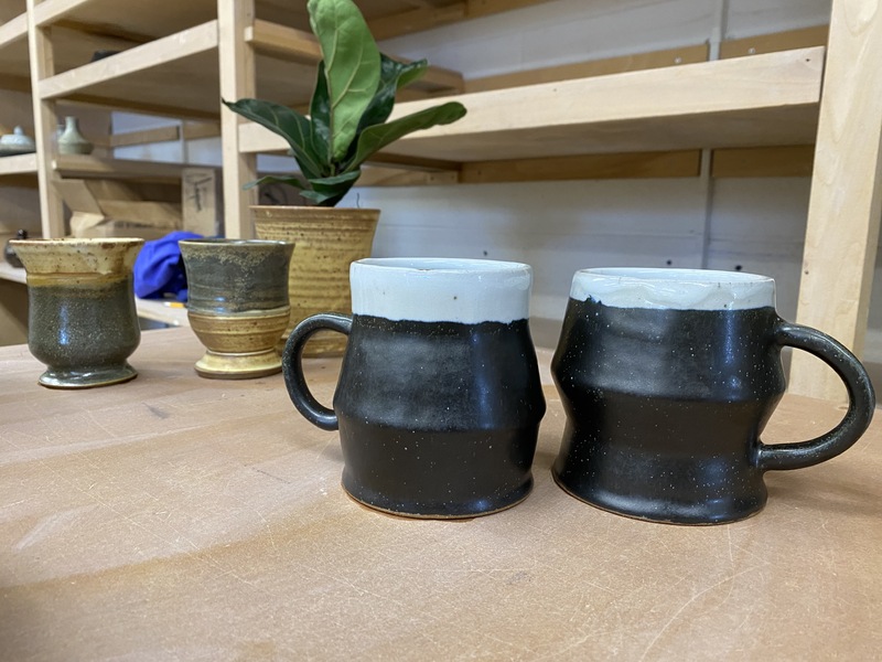 Four clay cups. In the front are two mugs that are black on the bottom and pale blue around the top edge and lip. Further back are two more cups, one with a green base and a tan top and the other with a tan base and a green top. In the background is a green plant in a tan pot and wooden shelves. 