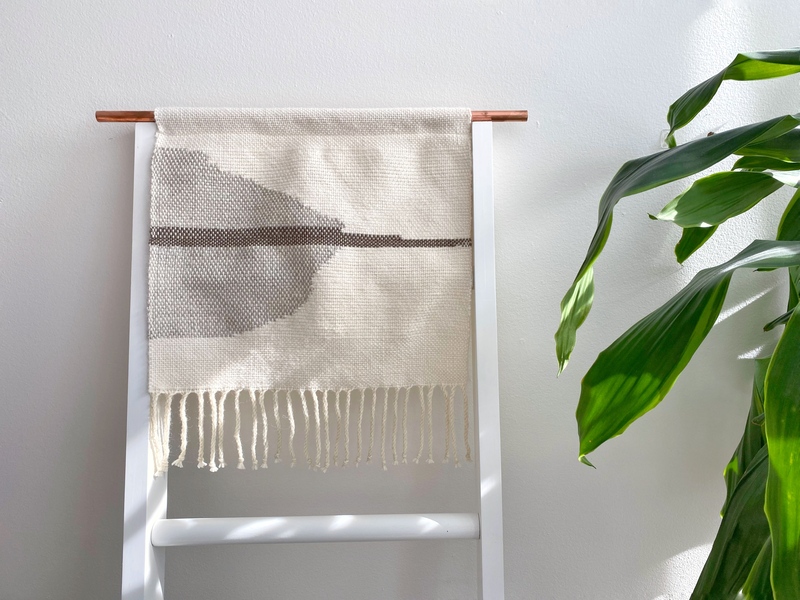 A photograph on a white backdrop. Draped over a white ladder in the center is a tapestry with tassels that reaches from the top rung to just above the third rung. The tapestry fabric is beige, with a dark brown-grey stripe running horizontally through the center and a lighter brown-grey organically shaped triangle that comes vertically from the left edge to the center. To the right are the long, tapering green leaves of a plant.