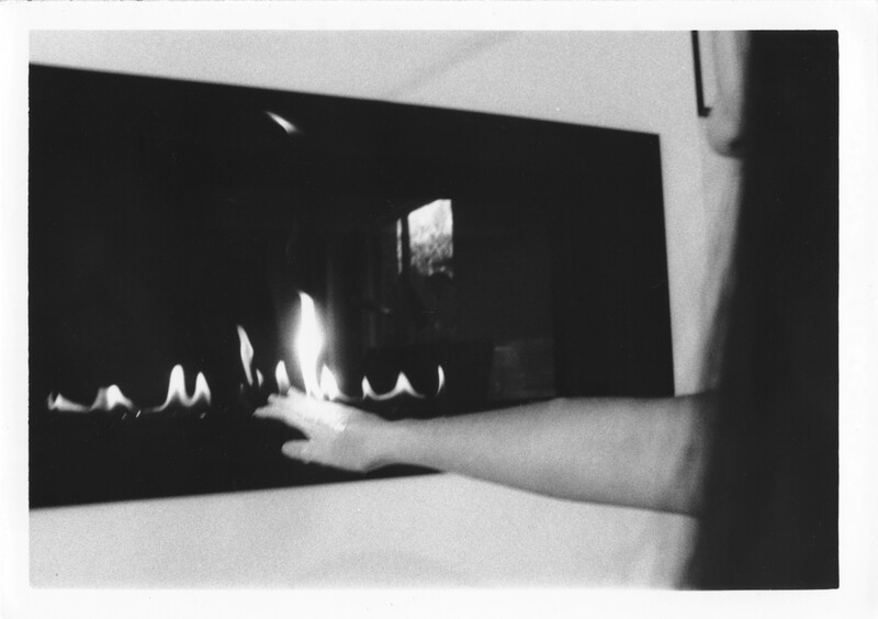 This is a sequence of images titled “Calcifer’s Curse” or A black and white film-processed photograph of a person reaching for a fireplace. The fireplace is black and set into a white wall with stark contrast. Flames rise from the base. From the right, someone is reaching their arm towards it. The flames hover above their outstretched fingers. The photograph is grainy.