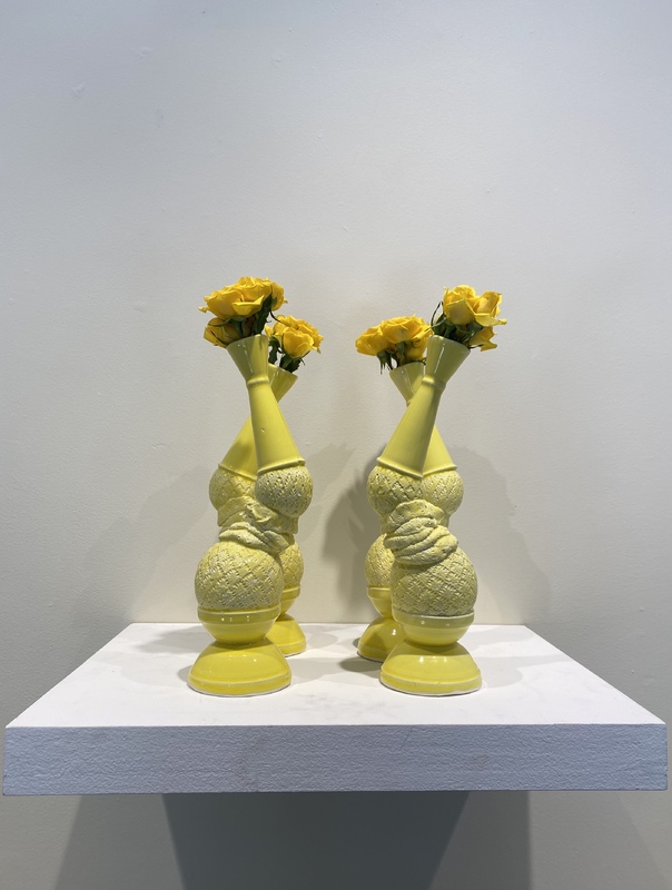 A photograph of four identical abstract vases, placed in two rows. The vases are yellow, with five distinct parts. The base of the vase is smooth and shaped like an upside-down bowl. Above the base is a round ball shape the size of an orange with a criss-cross pattern. Above that is a bunched-up section that connects to another round shape, this one the size of a lemon. On top of the lemon is an upside-down funnel shape with a right-side up funnel shape on top of it. Three yellow roses stick out the top of each base.