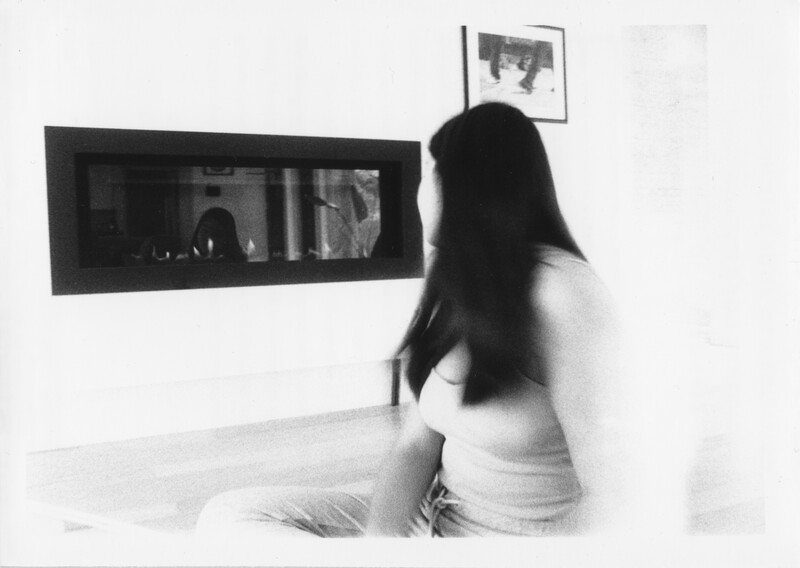 A black and white film processed photograph of a person in a brightly lit room. The person is light skinned with dark hair past their shoulders, shown from the waist up. They are looking over their right shoulder; their torso and left arm are blurred by the movement. Behind them against a white wall is a black built-in rectangular fireplace with small flames rising from its base. The glass on the fireplace reflects the person&#039;s head and the room. The photo is slightly grainy.