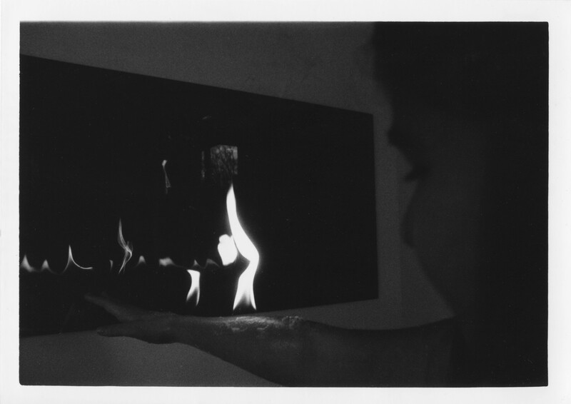 A dark black and white film-processed photograph of the silhouette of a person reaching towards a built-in fireplace. There is a stark contrast between the bright flames and the very dark surroundings. From the right, the silhouette of a person’s face is barely visible, and their arm is reaching towards the flames. The flames illuminate their forearm. The photograph has a grainy texture. 