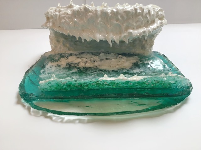 A photograph of a resin sculpture shaped like a cresting ocean wave, on a white backdrop, viewed vertically from the front with the wave cresting towards the viewer. The base of the sculpture is the semi-transparent blue-green of the ocean. White swirls coat the top of it like swirling sea foam. The wave is in the back, rising from the base and curling over in the shape of a letter C, with the top of the wave coming forward over the ocean base. 